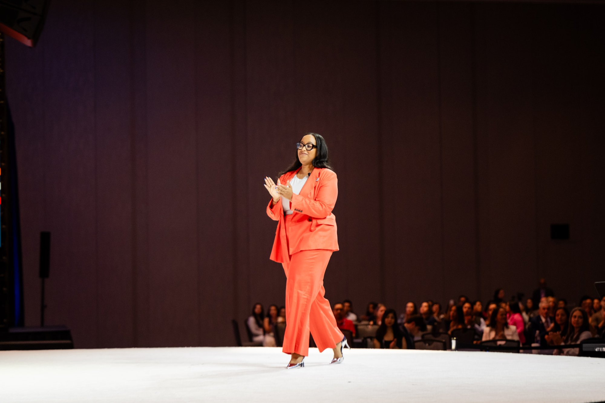 Jennylee Ramos on stage in a coral suit, mid-applause for the audience